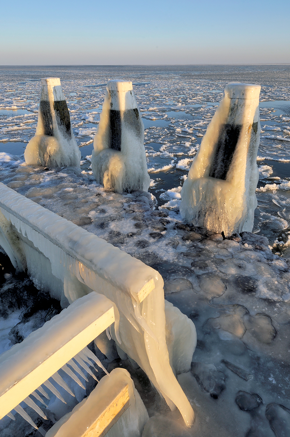 IJspegels hangen aan het hekwerk van een steiger met uitzicht op drijvende ijsschotsen tijdens winter op het IJsselmeer.