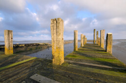 Zonlicht schijnt op de verweerde, houten palen en planken van een steiger in de haven van De Cocksdorp op het waddeneiland Texel.