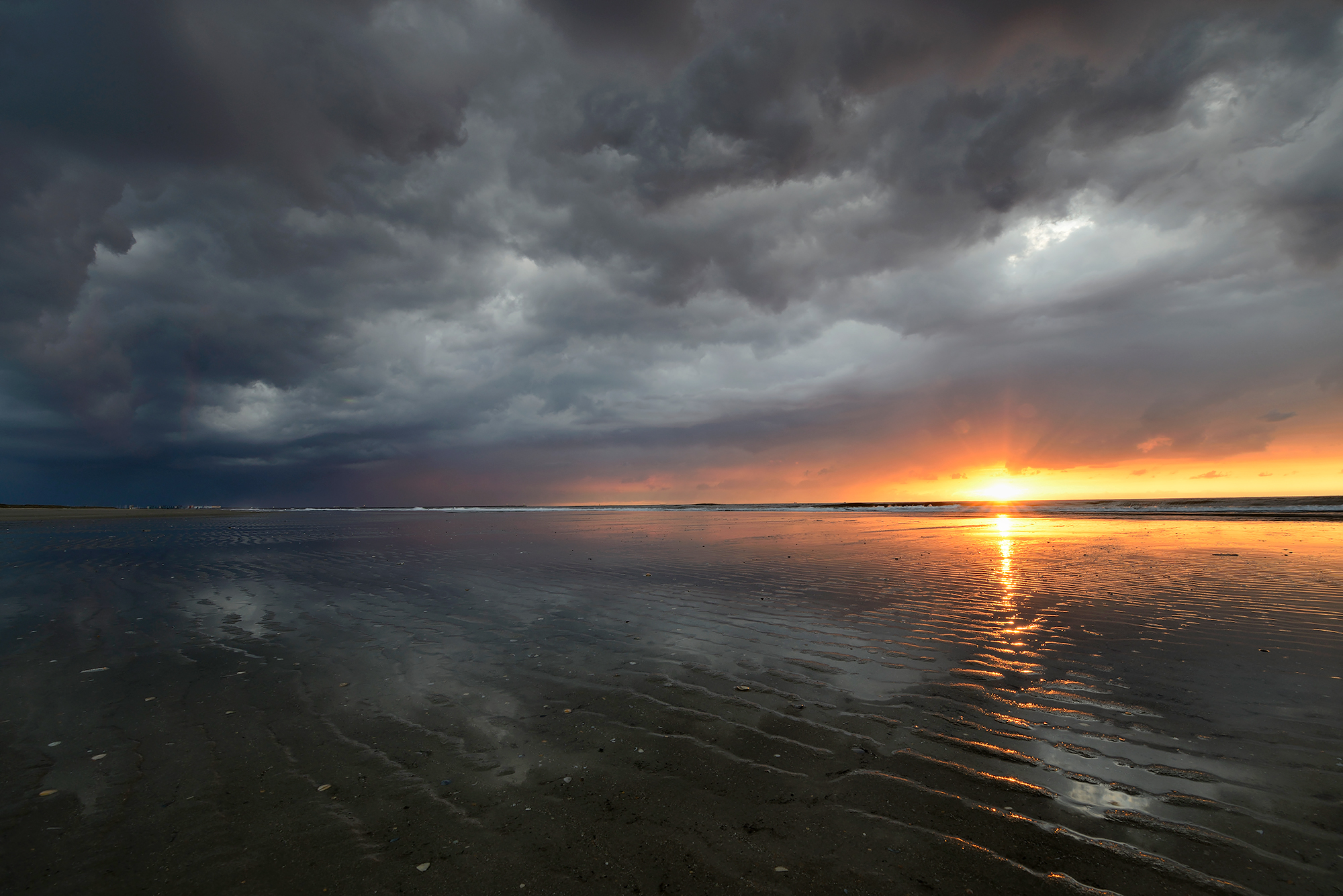 Laatste zonnestralen schijnen over het natte zand bij de vloedlijn tijdens zonsondergang met dreigende wolkenlucht op het strand van Wijk aan Zee