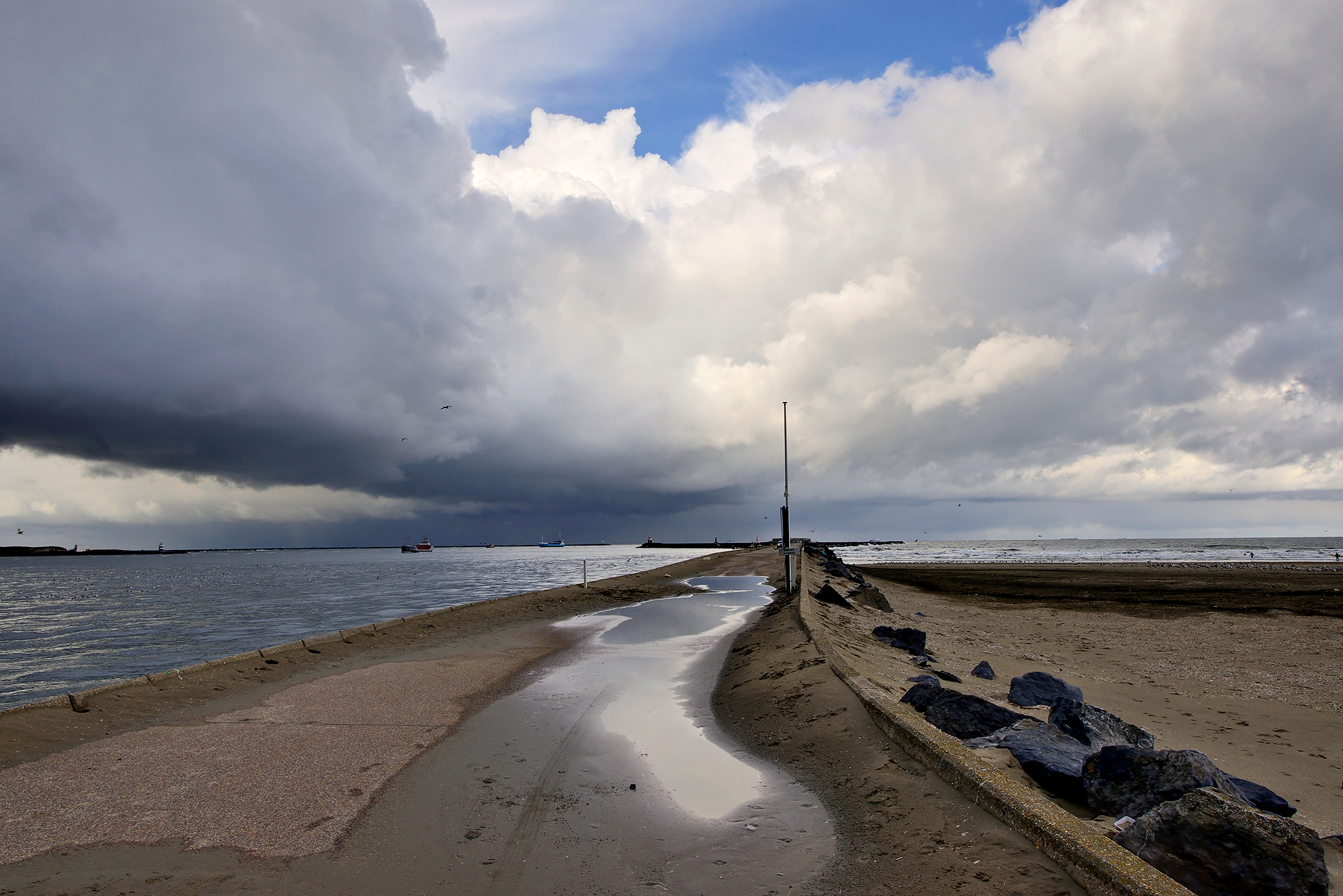 Donkere wolkenlucht van een regenbui strekt zich uit boven het strand en het betonnen pad van de Noordpier bij Wijk aan Zee.