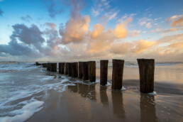 Weerspiegeling van warm zonlicht op wolken boven rij houten palen van paalhoofd tijdens zonsopkomst op het strand bij Schoorl