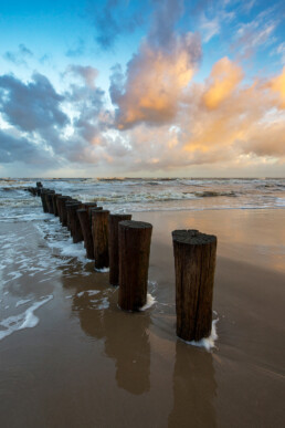 Weerspiegeling van warm zonlicht op wolken boven rij houten palen van paalhoofd tijdens zonsopkomst op het strand bij Schoorl