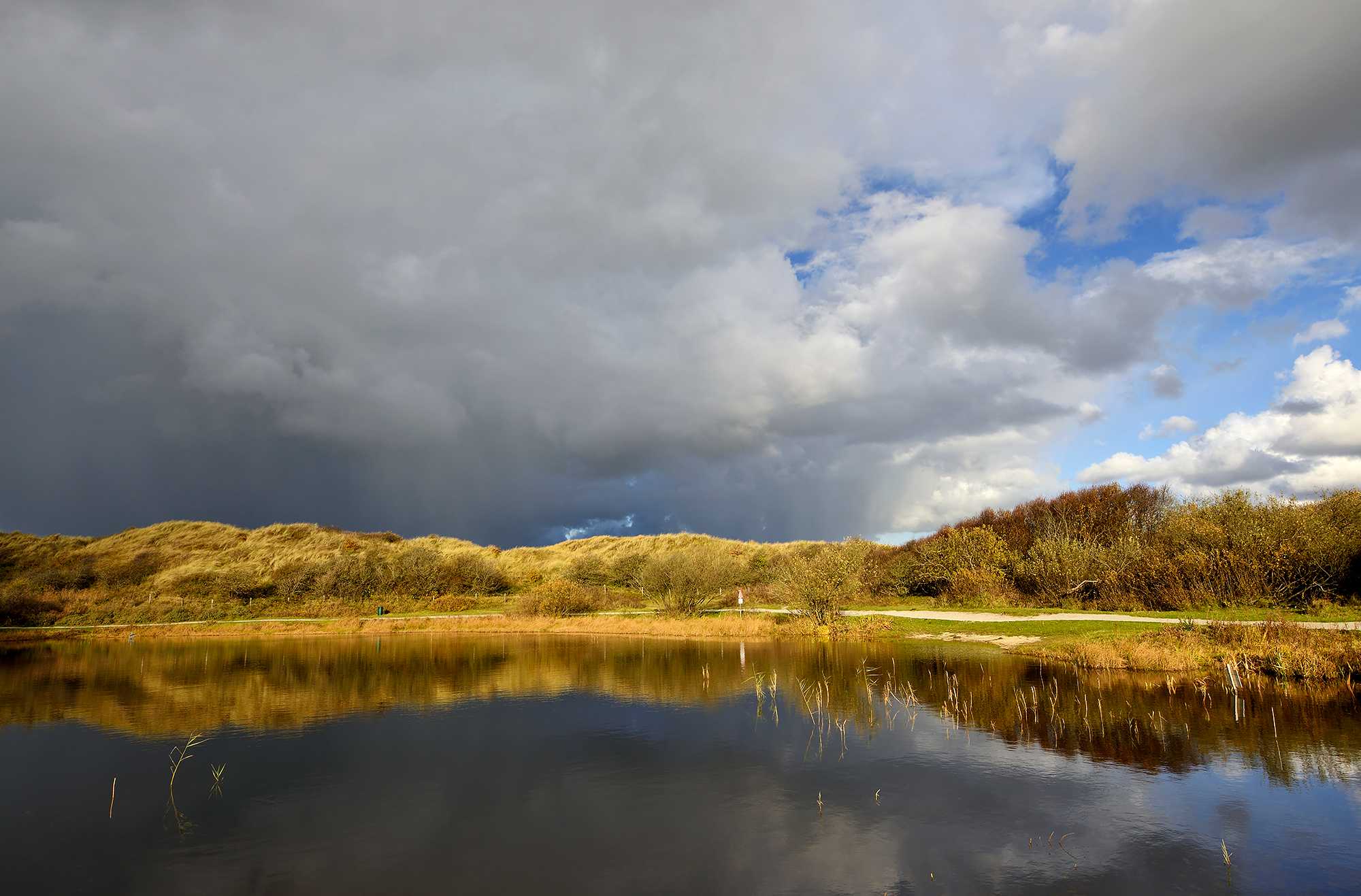 Donkere wolkenlucht van regenbui weerspiegelt in het stille water van een duinmeer in de Pettermerduinen bij Petten.