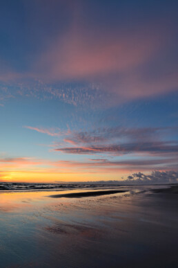 Warme gloed van zonsondergang weerspiegelt in het ondiepe zeewater langs de vloedlijn op het strand van Wijk aan Zee.