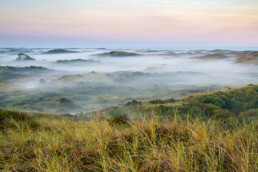 Uitzicht vanaf duintop over lagen mist tussen duinen tijdens zonsopkomst in het Noordhollands Duinreservaat bij Egmond aan Zee.